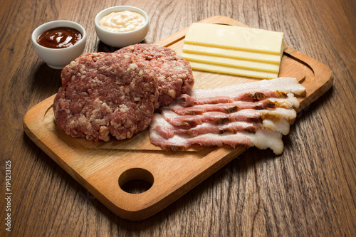 Ingredients for cooking burgers. Raw ground beef meat cutlets on wooden chopping board, cheese, bacon, over wooden background