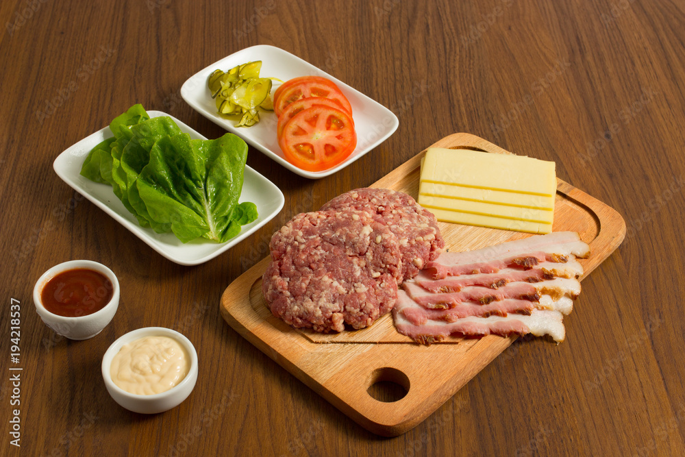 Ingredients for cooking burgers. Raw ground beef meat cutlets on wooden chopping board, tomatoes, greens, pickles, ketchup, cheese, over wooden background Ingredients for cooking burgers. Raw ground beef meat cutlets on wooden chopping board, tomatoes, greens, pickles, ketchup, cheese, over wooden background