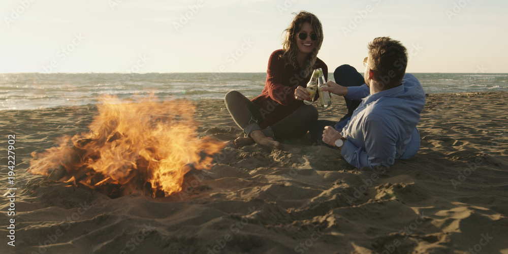 Loving Young Couple Sitting On The Beach beside Campfire drinking beer Loving Young Couple Sitting On The Beach beside Campfire drinking beer
