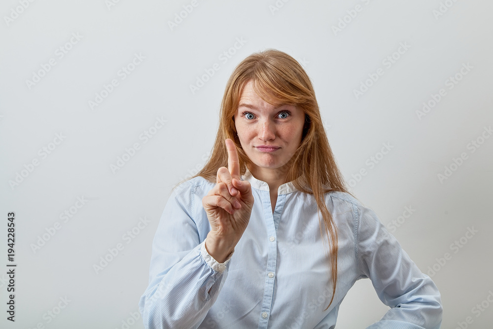 Headshot portrait of young european lady with red hair, white skin and freckles joyfully smiling and showing a note sign with her index finger raised, probably stopping somebody. Headshot portrait of young european lady with red hair, white skin and freckles joyfully smiling and showing a note sign with her index finger raised, probably stopping somebody.