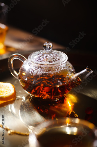 Small glass teapot and glasses with hot black tea, dried rose petals, pocket magnifier on golden chain, squeezed orange slice on golden tray. Evening light. Side view.