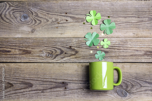 Green mug with four-leaf clover on wooden background. Copy space, top view.