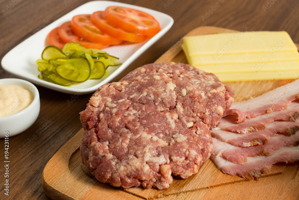 Ingredients for cooking burgers. Raw ground beef meat cutlets on wooden chopping board, tomatoes, greens, pickles, ketchup, cheese, over wooden background - Top View Ingredients for cooking burgers. Raw ground beef meat cutlets on wooden chopping board, tomatoes, greens, pickles, ketchup, cheese, over wooden background - Top View