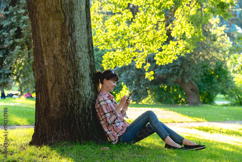 young woman sits under a tree with a phone in hands in the summe