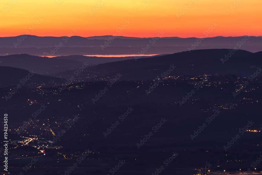 Beautiful aerial view of Umbria (Italy) valley at dusk, with hills, Perugia city lights and Trasimeno lake reflecting orange sky light