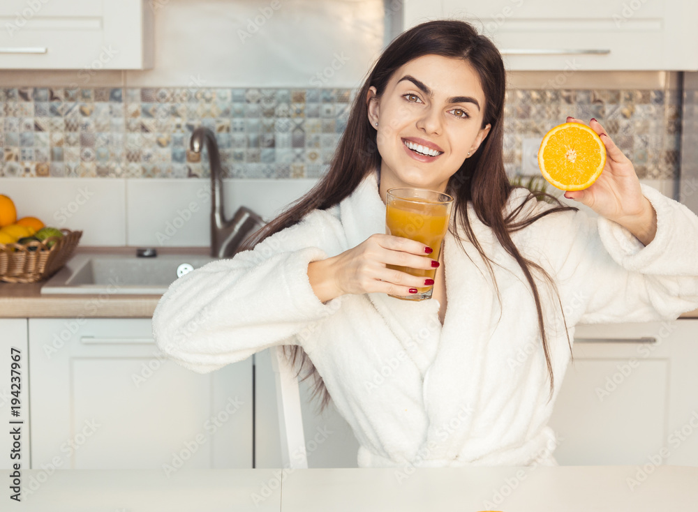 Beautiful girl wears white bathrobe with glass of orange juice and fruit, kitchen morning sunset view Beautiful girl wears white bathrobe with glass of orange juice and fruit, kitchen morning sunset view