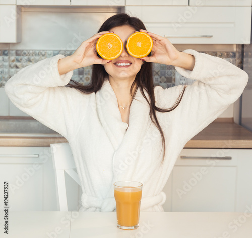 Beautiful girl wears white bathrobe with glass of orange juice and fruit, kitchen morning sunset view