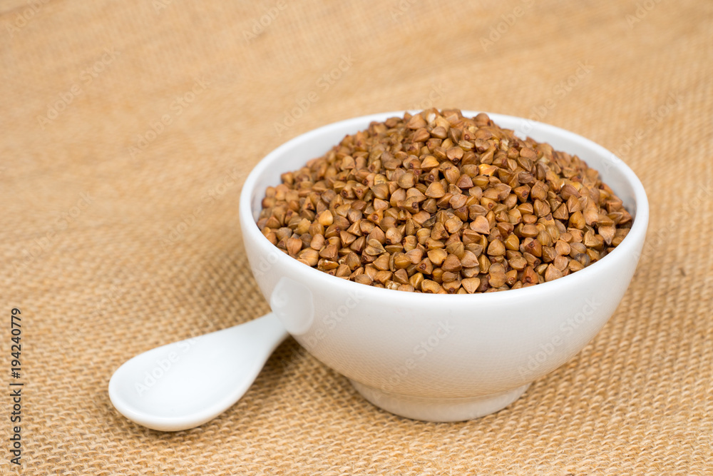 dry buckwheat heap in white bowl with spoon over sackcloth dry buckwheat heap in white bowl with spoon over sackcloth