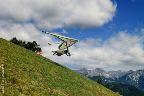 Pilot runs with a hang glider on a green grassy slope high in the mountains with blue sky and clouds above