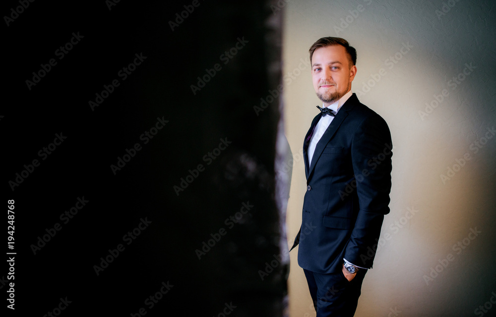 Handsome groom in classy black suit stands in a dark room Handsome groom in classy black suit stands in a dark room