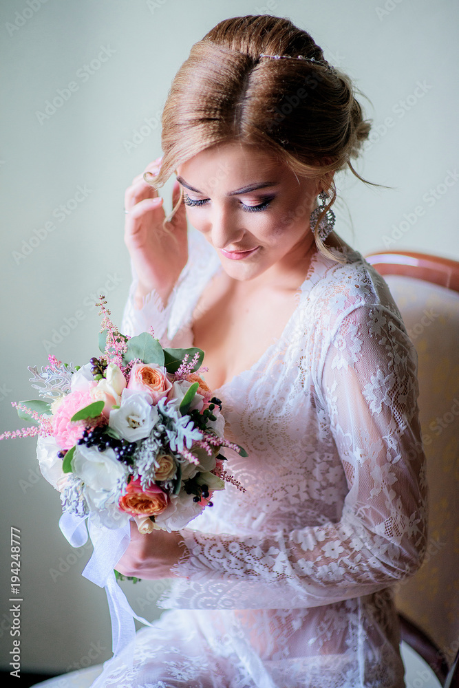 Charming blonde bride in sexy white robe sits with wedding bouquet on the chair Charming blonde bride in sexy white robe sits with wedding bouquet on the chair