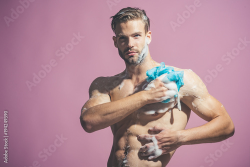 Attractive stylish macho model with fashion hair takes shower with soap sponge. Bare man is washed chest with soap sponge in bathroom. Man with bristle and concentrated face take shower. Copy space.