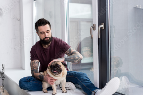 tattooed handsome man sitting on windowsill and palming dog at home