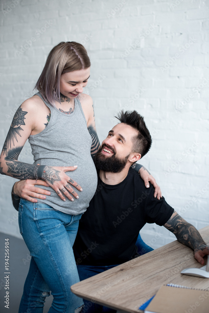 smiling tattooed boyfriend hugging pregnant girlfriend at home