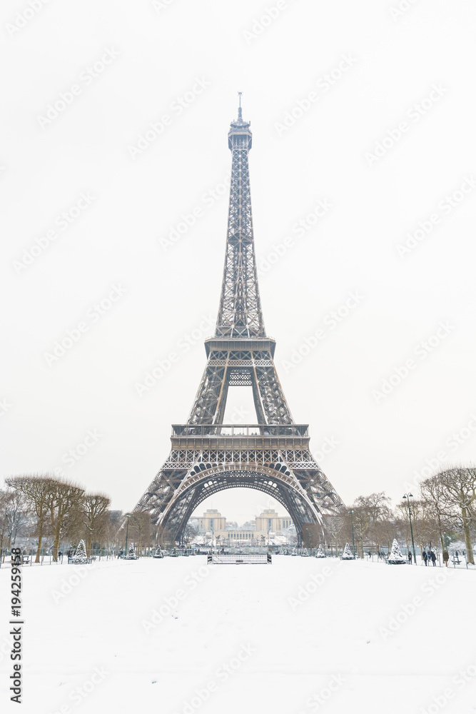 Winter in Paris in the snow. The Eiffel tower seen from the Champ de Mars, with a snow covered lawn in the foreground and the top of the tower disappearing slightly in the mist.