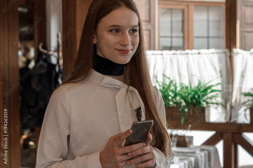 Fototapeta premium a young girl with straight long hair and brown eyes in the restaurant holds the phone in her hands and thinks what message to write, smiles and looks aside