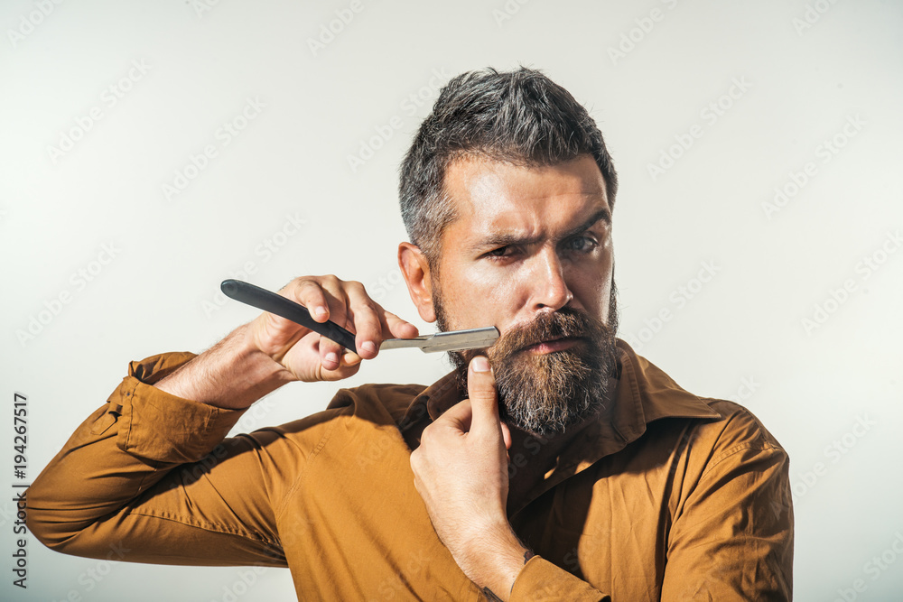 Obraz premium Professional barber with thick beard and mustache, holding straight razor. Hairdresser demonstrating sharp blade of barbershop accessory. Young bearded macho shaving with razor blade.