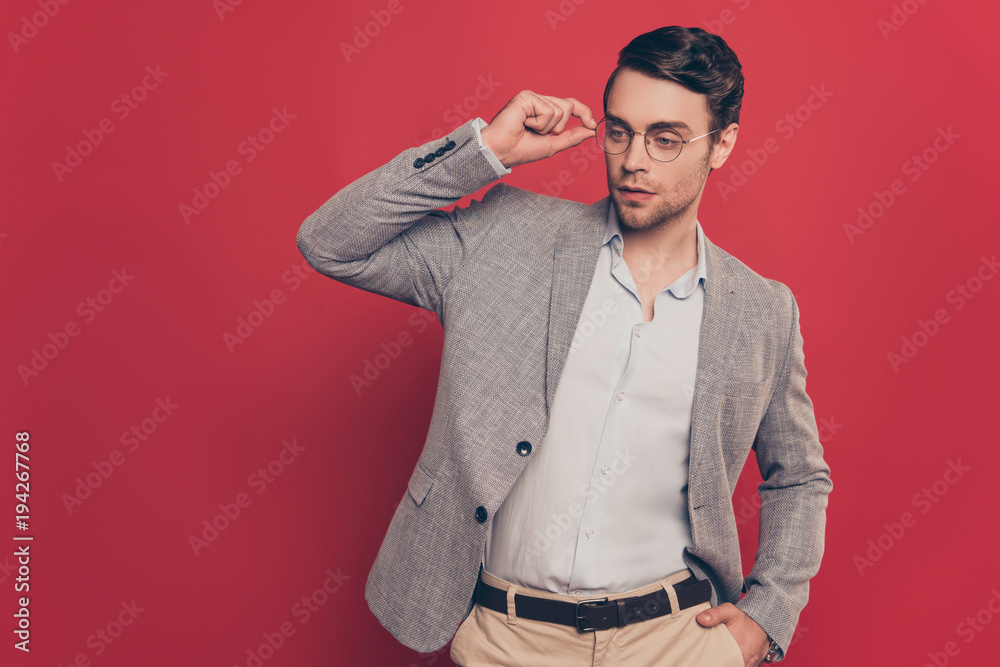 Portrait of stunning, virile, harsh, modern man holding hand in pocket of pants and eyelet of glasses on his face, looking to the side, standing over red background