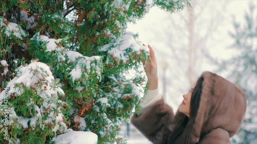 Close-up of brunette stylish girl with bright makeup, near Snow covered Christmas trees, in a luxurious brown fur coat walking in winter time slow motion