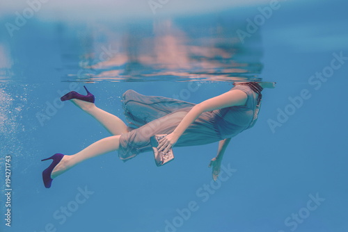 surreal art portrait of young woman in grey dress, beaded scarf , small bag, violet high heels underwater in the swimming pool 