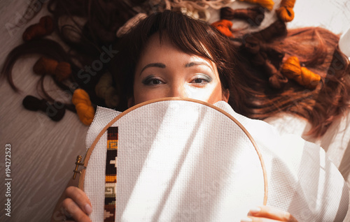 Young beautiful woman is engaged in embroidering a dagger on the embroidery frame in a colored dress at the window in the sunlight. A great picture for a hobby