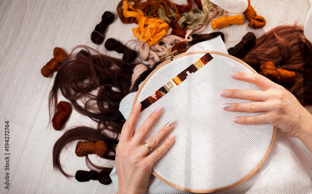 Φωτογραφία Young beautiful woman is engaged in embroidering a dagger on the embroidery frame in a colored dress at the window in the sunlight