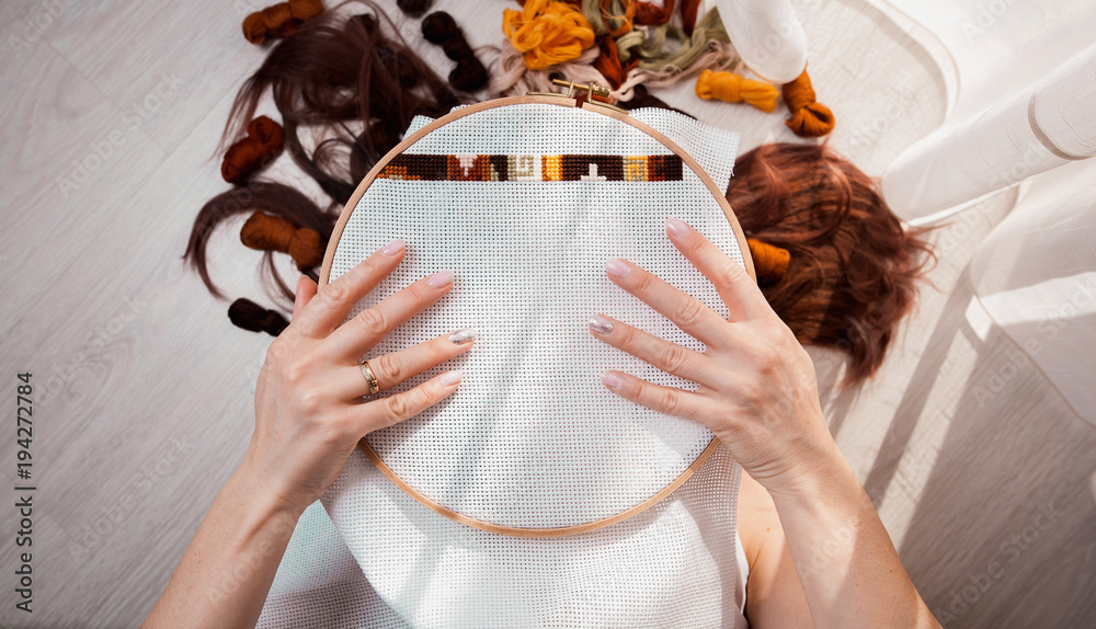 Φωτογραφία Young beautiful woman is engaged in embroidering a dagger on the embroidery frame in a colored dress at the window in the sunlight