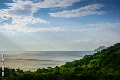 Evening sea landscape with sun rays and green wooded coast. 