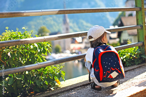 a small child with a backpack sits with his back and admires the cityscape in the town of Hallstatt Austria