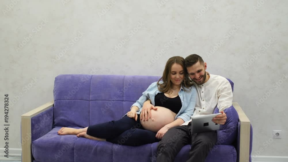 Happy family couple sitting on the couch calling on video communication to their parents in another city. Tells news about pregnancy. A pregnant woman and a man communicate via a tablet computer via Happy family couple sitting on the couch calling on video communication to their parents in another city. Tells news about pregnancy. A pregnant woman and a man communicate via a tablet computer via