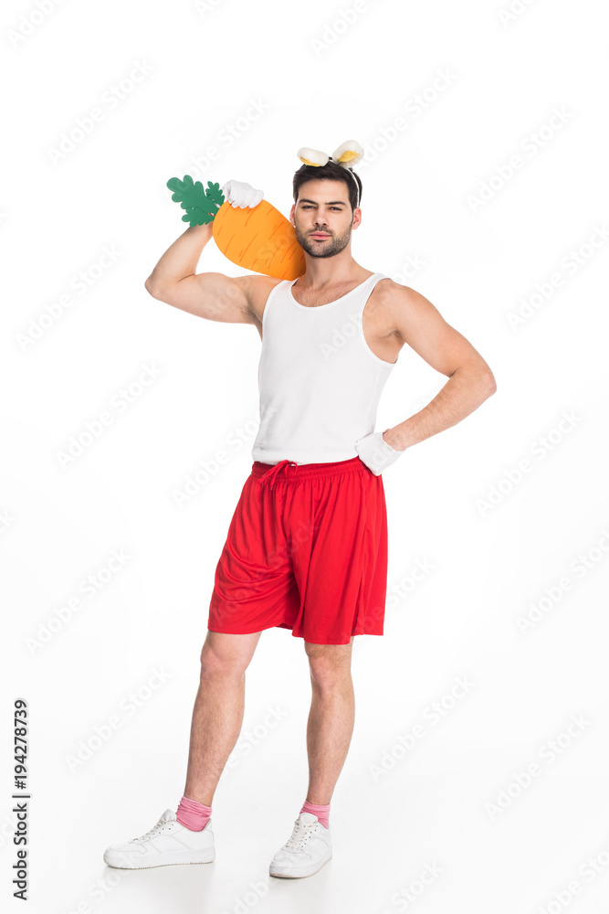 Naklejka premium View of man with bunny ears holding big carrot on shoulder isolated on white, easter concept