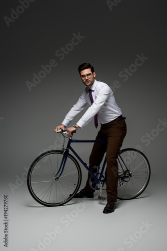  stylish businessman in glasses sitting on bicycle on grey