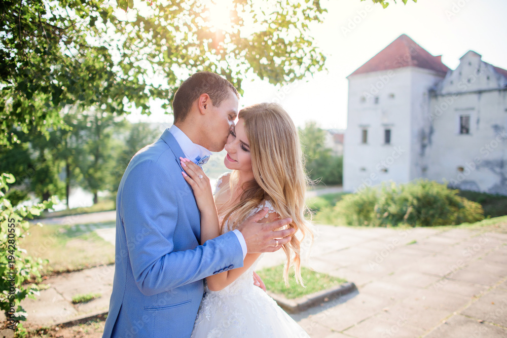 Groom holds bride tender posing with her before an old castle in a sunny summer day