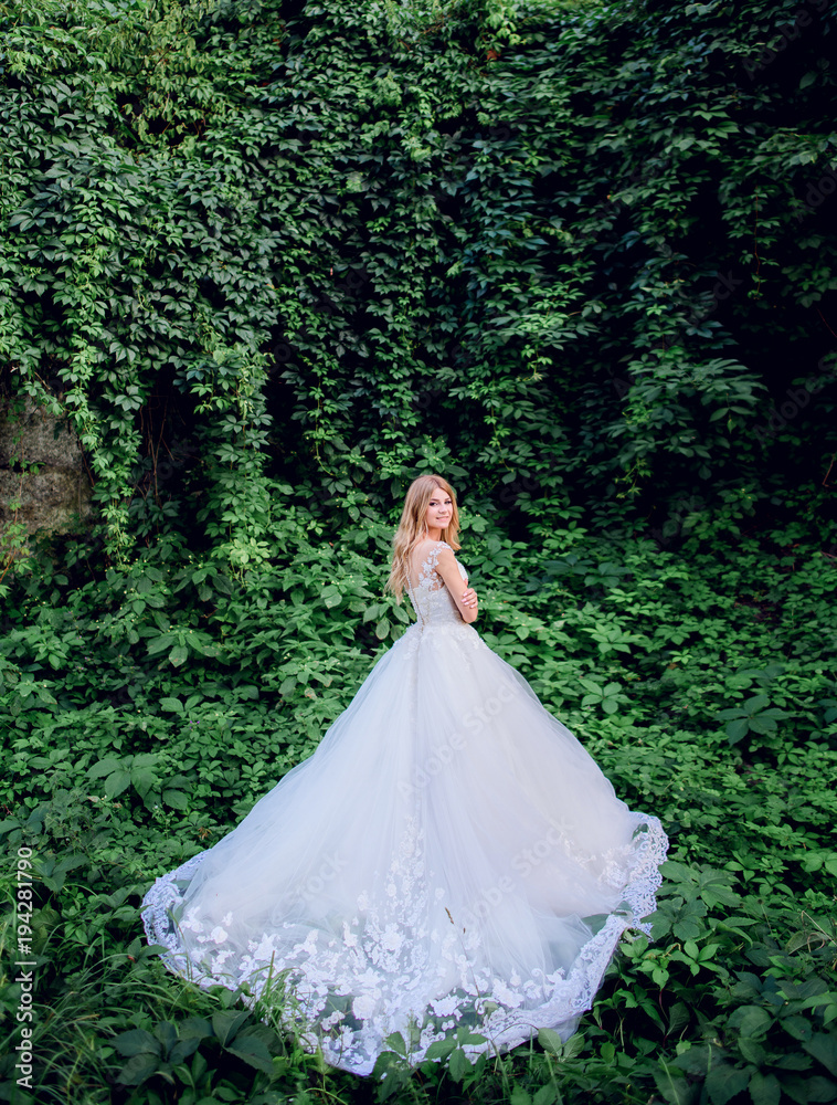 Blonde bride in rich wedding dress poses among greenery outside