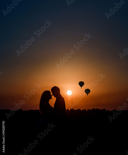 Silhouettes of a couple hugging each other before a sunset and air balloons up in the sky