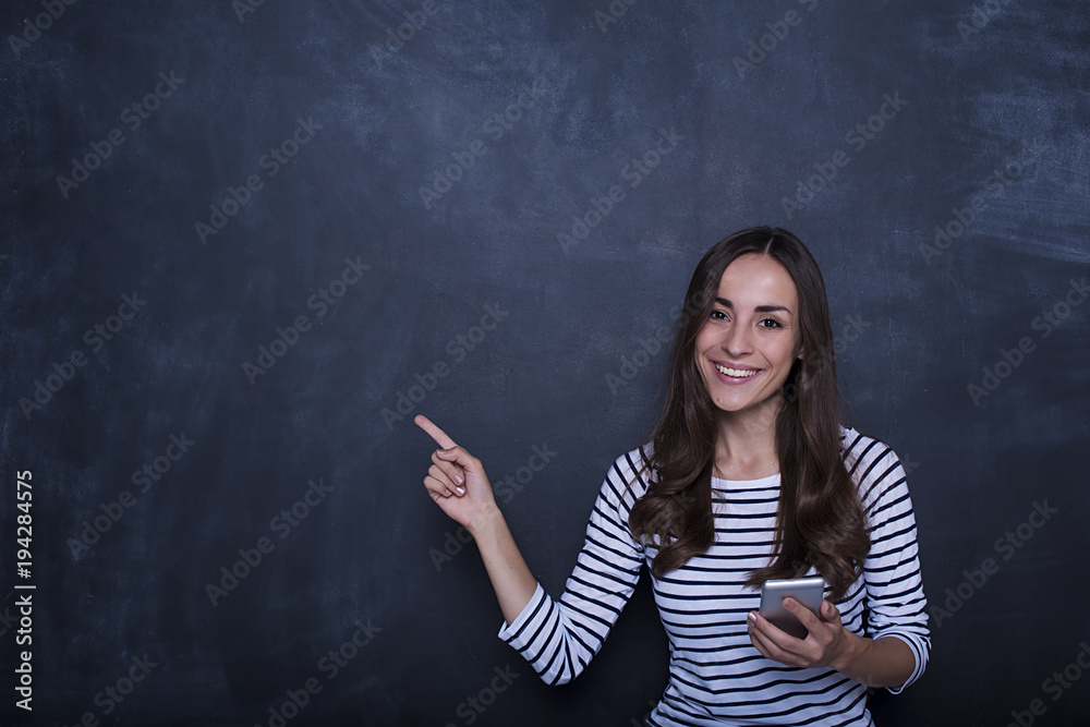 Pretty happy young woman holding using smartphone standing over chalkboard background and pointing on copy space to the side