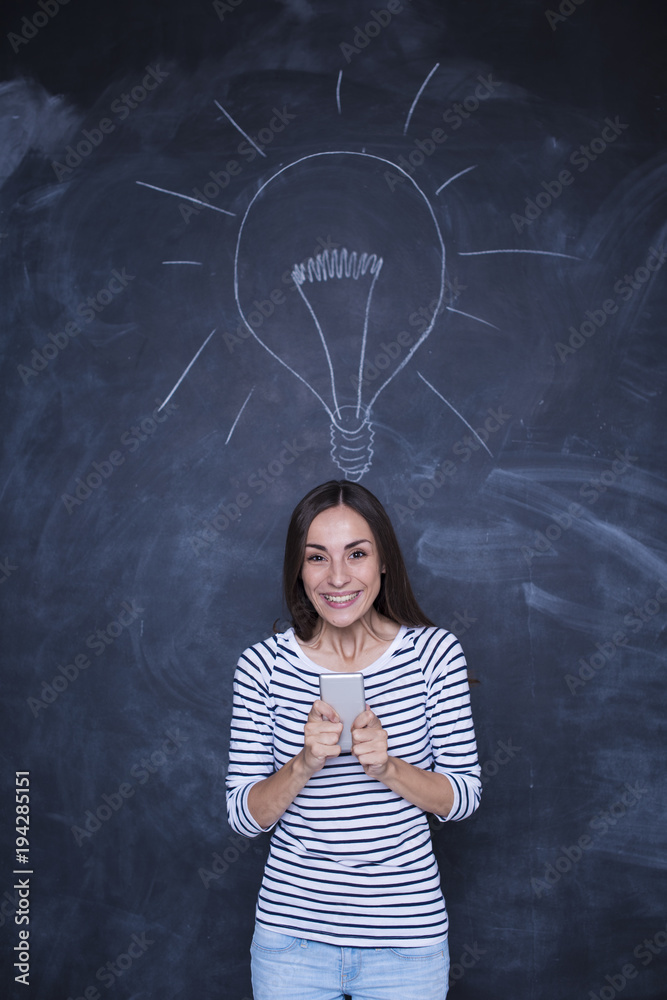 Happy young woman holding smartphone standing over chalkboard background with light bulb drawn