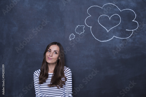 Attractive thoughtful young woman at the blackboard with a drawn cloud and heart over her head.