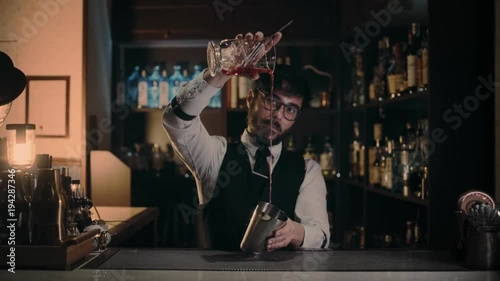 Professional bartender in dress suit is pouring cooled down rose vermouth from glass with ice cubes to stailess steel shaker in private hidden secret bar, looking seriously in camera.Dark indoor light photo