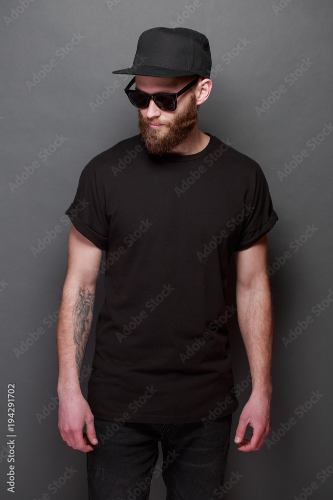 Hipster handsome male model with beard wearing black blank t-shirt with space for your logo or design over gray background