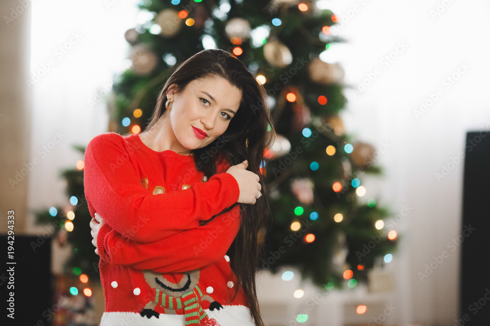 Pretty young woman with long brinette hair poses in red sweater before a Christmas tree Pretty young woman with long brinette hair poses in red sweater before a Christmas tree