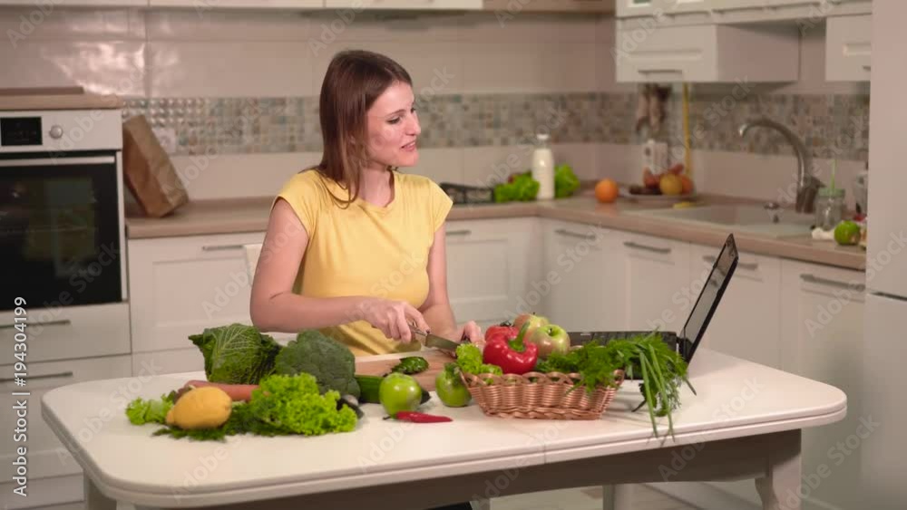 Sweet fair-haired girl in yellow t-shirt slicing cucumber, talking with pleasure over the laptop during meal prep, indoor shot in furnished light kitchen Sweet fair-haired girl in yellow t-shirt slicing cucumber, talking with pleasure over the laptop during meal prep, indoor shot in furnished light kitchen