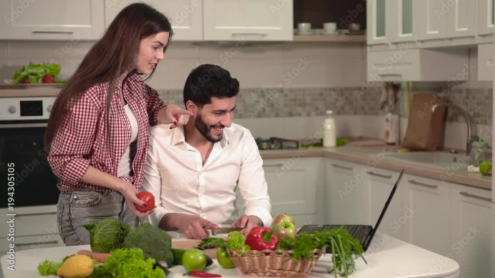 Handsome dark-haired man in neat white shirt slicing cucumber, cheerful beautiful woman in red shirt pointing at the laptop, having lovely evening talk in cozy modern kitchen Handsome dark-haired man in neat white shirt slicing cucumber, cheerful beautiful woman in red shirt pointing at the laptop, having lovely evening talk in cozy modern kitchen