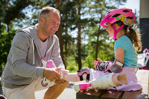 Father helping his daughter in helmet to wear protective pads for roller skates. Happy family spending time together. Sunny summer day on suburb street photo