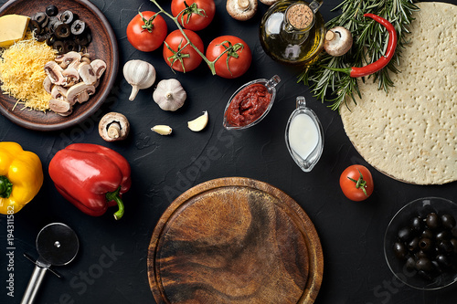 Cutting wooden board with traditional pizza preparation ingredients: cheese, tomatoes sauce, basil, olive oil, pepper, spices. Black texture table background