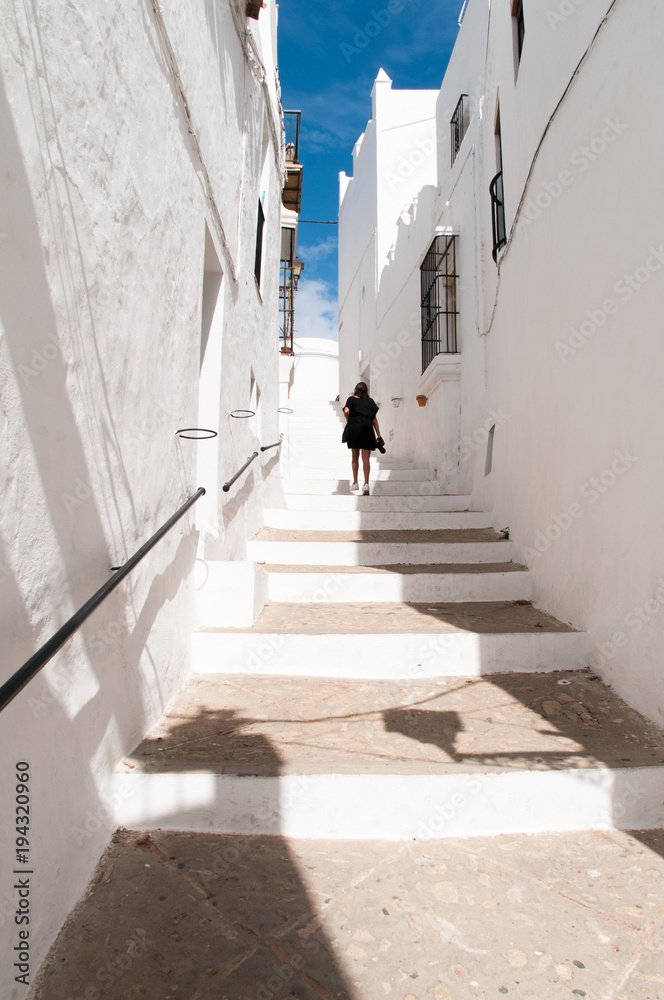 Female tourist on black dress and camera in hand walks upstairs on traditional narrow street in Vejer white town in Andalusia, Spain