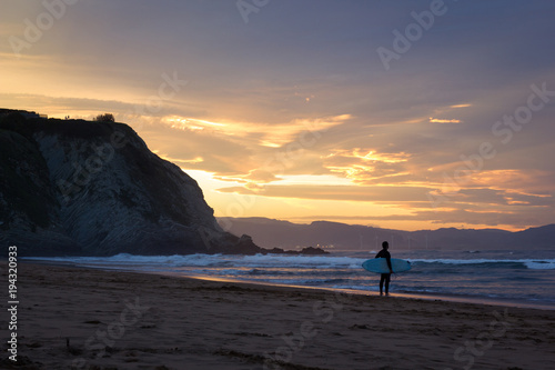 Lonely surfer with long black wet suit and surfing board on hand stands on shore at splendid sunset in Arrietara beach, north Spain. Man contemplating waves waiting for his set to surf photo