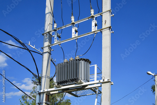 An electrical distribution transformer with cooling fins is located on the pole. Against the blue sky.