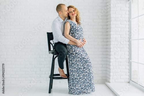 Man sitting barefoot on high chair hugging his pretty pregnant wife in long dress.