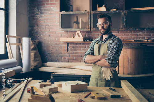 Serious concentrated confident qualified handsome bearded cabinet-maker wearing apron and safety glasses is standing with crossed arms in his workshop and going to work with wood